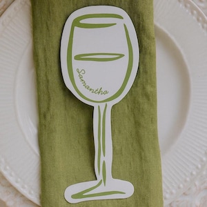 Könnte beinhalten: Eine weiße Tischkarte in Form eines Weinglases mit dem Namen "Samantha" in grüner Schrift. Die Karte liegt auf einer grünen Serviette, die auf einem weißen Teller liegt. Das Weinglas-Design hat grüne Umrisse.
