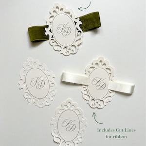 May include: Four white oval paper emblems with intricate die-cut borders and the monogram KD in script. One emblem features a green velvet ribbon, another a white satin ribbon, and two are plain. Text labels read Belly Band Emblem and Includes Cut Lines for ribbon.