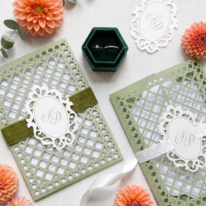 May include: Two sage green laser-cut gatefold wedding invitations with white inserts, featuring an oval monogram tag with the letters KD, secured by velvet and satin ribbons, alongside a dark green hexagonal ring box with two rings.