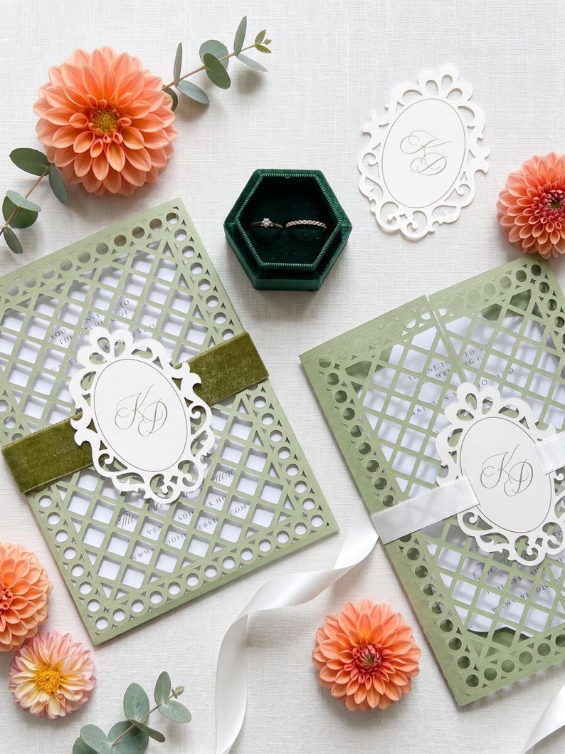 May include: Two sage green laser-cut gatefold wedding invitations with white inserts, featuring an oval monogram tag with the letters KD, secured by velvet and satin ribbons, alongside a dark green hexagonal ring box with two rings.