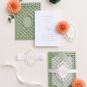 May include: A wedding invitation suite featuring a white card with black text, a sage green laser-cut lattice folder, a white ribbon belly band with an oval monogram tag, peach dahlia flowers, and a dark green velvet hexagonal ring box.