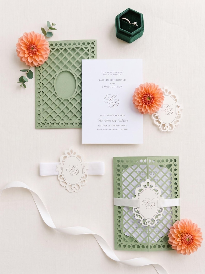 May include: A wedding invitation suite featuring a white card with black text, a sage green laser-cut lattice folder, a white ribbon belly band with an oval monogram tag, peach dahlia flowers, and a dark green velvet hexagonal ring box.