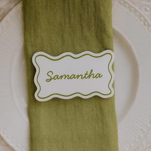 May include: A white place card with a scalloped edge and the name "Samantha" in green script. The place card rests on a folded green napkin, with a white plate in the background.