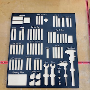 Op de afbeelding: Een zwarte gereedschapsorganizer met uitsparingen voor verschillende gereedschappen, waaronder schuifmaten, sleutels en boortjes. De organizer heeft labels voor elke uitsparing, zoals "1/4 Up-Cut", "1/4V Bits", "Calipers" en "Locating Pins".