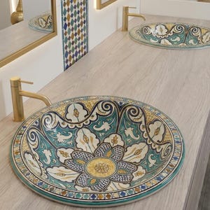 Puede incluir: Un mueble de baño con dos lavabos decorativos. Los lavabos son redondos y ovalados, con un diseño floral en verde azulado, amarillo y blanco. Grifos y espejos dorados complementan los lavabos. El mueble tiene un acabado de madera clara y las paredes son blancas.