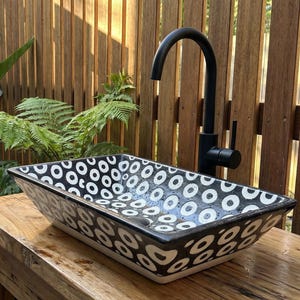 Könnte beinhalten: Ein rechteckiges Keramikwaschbecken mit einem schwarz-weißen Muster aus Kreisen und Ovalen. Das Waschbecken ist auf einem Holztisch montiert, mit einem schwarzen Wasserhahn. Der Hintergrund zeigt einen Holzzaun und üppiges Grün.
