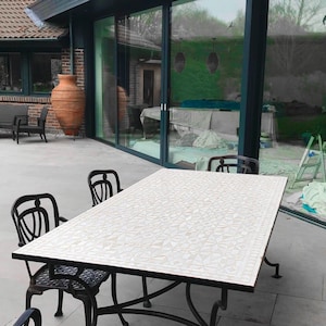 Puede incluir: Un juego de comedor al aire libre con una mesa rectangular con tapa blanca estampada y patas de metal negro. Cuatro sillas de metal negro con respaldos ornamentados rodean la mesa. El conjunto está sobre un patio de piedra.