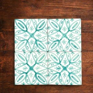 Peut inclure: Quatre carreaux de céramique carrés avec un motif floral turquoise et blanc disposés sur une surface en bois. Les carreaux présentent un motif symétrique avec des feuilles et des pétales stylisés, créant un aspect décoratif et artistique.