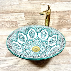 Op de afbeelding: Een ovaal keramische wastafel met een turquoise en wit bloemenpatroon. De wastafel heeft een goudkleurige afvoer en wordt gecombineerd met een gouden kraan. De rand van de wastafel is effen turquoise en het ontwerp is gecentreerd.