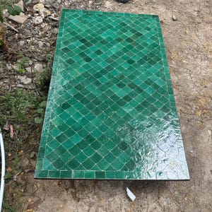 May include: Rectangular green tile table with a diamond pattern. The tiles are a vibrant shade of green, arranged in a repeating pattern. The table has a dark border and is set outdoors.
