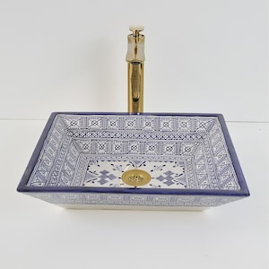 Op de afbeelding: Een rechthoekige keramische wastafel met een blauw-wit geometrisch patroon. De wastafel heeft een gouden kraan en afvoer. De wastafel is een opzetmodel, ontworpen om op een wastafelblad te worden geplaatst.