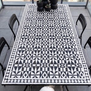 May include: A rectangular outdoor dining table with a black and white geometric tile pattern. Four black metal chairs surround the table. Small potted plants sit on the table. A small white dog sits under the table.
