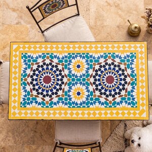 May include: A rectangular mosaic tile table with a yellow border and intricate geometric patterns in blue, teal, red, and white. The table is surrounded by wrought iron chairs with cream-colored cushions. A small dog rests on a rug nearby.