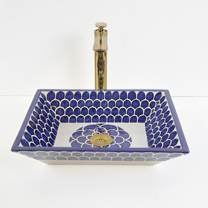 Op de afbeelding: Een rechthoekige keramische wastafel met een blauw-wit patroon. De wastafel heeft een gouden kraan en een decoratieve afvoer. De binnenkant van de wastafel heeft een bloemenontwerp in het midden. Het is een decoratief badkameraccessoire.