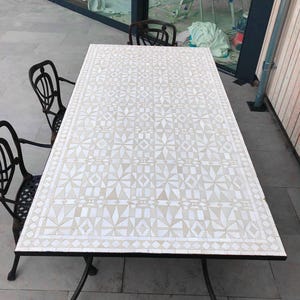 May include: A rectangular outdoor dining table with a mosaic tile top featuring a geometric pattern in shades of white and beige. The table has a black metal frame and is surrounded by black metal chairs.