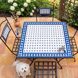 Könnte beinhalten: Garten-Terrassenset mit einem quadratischen Tisch und vier Stühlen. Der Tisch hat eine weiße Oberfläche mit blauen rautenförmigen Fliesen. Die Stühle haben passende blaue und weiße Fliesenakzente. Ein kleiner weißer Hund sitzt in der Nähe des Tisches.