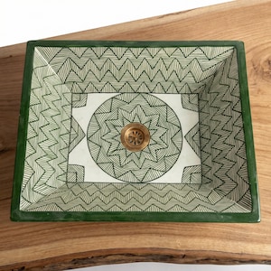 Op de afbeelding: Een rechthoekige keramische wastafel met een groene rand en een wit interieur versierd met groene geometrische patronen. De wastafel heeft een cirkelvormig ontwerp rond de afvoer en een messing afvoerdeksel. De wastafel staat op een houten oppervlak.