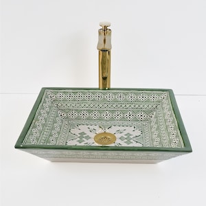 Op de afbeelding: Een rechthoekige keramische wastafel met een groen en wit geometrisch patroon. De wastafel heeft een gouden kraan en afvoer. De wastafel is een opzetmodel, ontworpen om op een wastafelmeubel te staan.