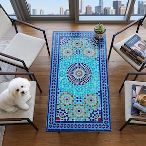 Op de afbeelding: Een rechthoekige mozaïektegel salontafel met een levendig turquoise, blauw en bruin bloemenpatroon. De tafel is omgeven door twee stoelen met lichtgekleurde kussens en donkere metalen frames. Een kleine witte hond zit op een stoel.