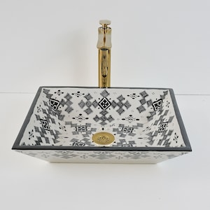 Op de afbeelding: Een vierkante, witte keramische badkamer wastafel met een zwart-wit geometrisch patroon. De wastafel heeft een goudkleurige kraan en afvoer. De rand van de wastafel is zwart. De wastafel is een vessel-stijl, ontworpen om op een aanrecht te staan.