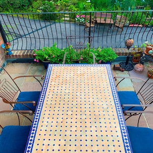 May include: Outdoor patio set featuring a rectangular table with a mosaic tile top in beige and blue. The set includes two metal chairs with blue cushions. The background shows a garden with lush greenery and a black wrought iron railing.