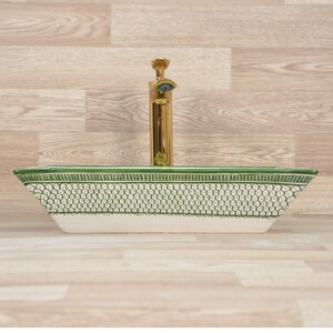 K&ouml;nnte beinhalten: Rechteckiges Keramikwaschbecken mit gr&uuml;n-wei&szlig;em Muster, kombiniert mit einer goldenen Armatur. Das Design des Waschbeckens weist ein detailliertes, sich wiederholendes Muster am oberen Rand und an den Seiten auf, das ein dekoratives Element bildet. Die Armatur hat ein schlankes, modernes Design.