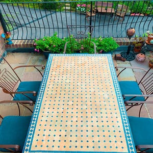 Peut inclure: Ensemble de patio extérieur avec une table rectangulaire ornée d'un dessus en mosaïque beige et bleu. Quatre chaises en métal avec coussins turquoise entourent la table. Le cadre est un jardin avec une végétation luxuriante et une clôture en fer forgé.