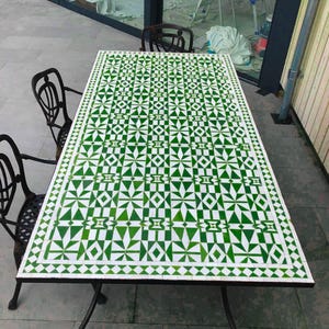 May include: A rectangular outdoor dining table with a white and green geometric tile top. The table is supported by black wrought iron legs and is surrounded by black wrought iron chairs. The table's surface features a repeating pattern of green shapes.