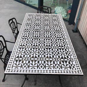 May include: Rectangular outdoor dining table with a black and white geometric tile top. The table has a black metal frame and is surrounded by black metal chairs. The tile pattern features triangles, diamonds, and starburst shapes.
