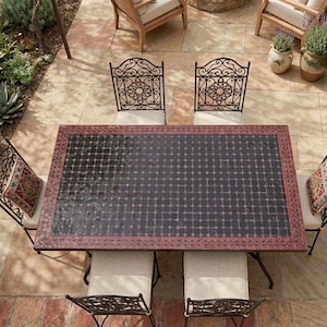 May include: Overhead shot of an outdoor dining set. The set features a rectangular table with a black tiled surface and a red border, surrounded by six wrought-iron chairs with cream cushions. Decorative cushions and potted plants complete the scene.