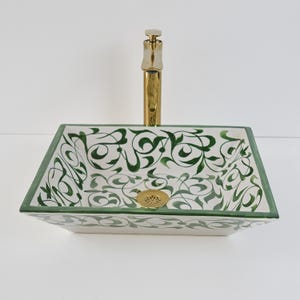 Op de afbeelding: Rechthoekige keramische wastafel met een witte basis en een groen bloemmotief. De wastafel heeft een goudkleurige kraan en afvoer. De randen van de wastafel zijn groen omlijnd. Een decoratief badkamerarmatuur.