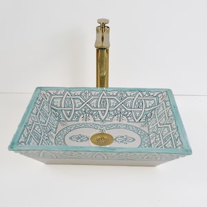 Op de afbeelding: Rechthoekige keramische badkamer wastafel met een gouden kraan. De wastafel heeft een witte basis met ingewikkelde turquoise en zwarte geometrische patronen. De rand is omlijnd in turquoise. Een gouden afvoer is zichtbaar.