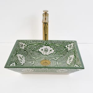 Op de afbeelding: Een vierkante keramische badkamer wastafel met een groen en wit bloemenpatroon. De wastafel heeft een gouden kraan en afvoer. De wastafel is een opzetmodel, met een rechthoekige vorm en een decoratief ontwerp.