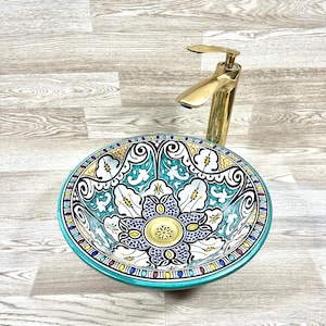 Pode incluir: Uma pia de cerâmica com um design marroquino colorido. A pia é redonda e tem um fundo branco com detalhes azuis, verdes, amarelos e vermelhos. A pia é decorada com padrões florais intrincados. Uma torneira dourada está montada na pia.