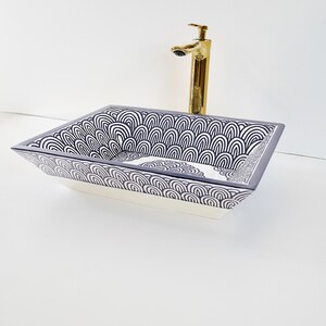 Op de afbeelding: Rechthoekige keramische wastafel met een witte basis en een decoratieve bovenkant met een zwart-wit golfpatroon. Een goudkleurige kraan is op het witte oppervlak achter de wastafel gemonteerd.