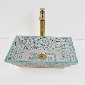 Op de afbeelding: Rechthoekige keramische badkamer wastafel met een gouden kraan. De wastafel heeft een witte basis met een turquoise rand en turquoise en zwarte bloemenkrullen. De afvoer is goudkleurig.