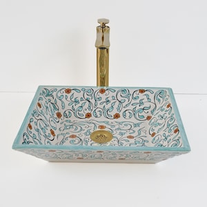 Op de afbeelding: Rechthoekige keramische badkamer wastafel met een bloemenpatroon in teal, oranje en zwart op een witte achtergrond. De wastafel heeft een teal rand en een gouden kraan en afvoer.