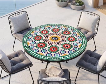 Op de afbeelding: Een ronde mozaïektegel tuintafel met een zwart metalen frame, omringd door vier draadstoelen met grijze kussens en kussens. Het tafelblad heeft een kleurrijk geometrisch patroon. Vetplanten in betonnen potten zijn op de achtergrond te zien.