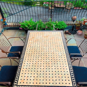 May include: A patio set with a rectangular table featuring a mosaic tile top in beige and black. The table is surrounded by four metal chairs with blue cushions. The background shows a garden with lush greenery and a wrought iron fence.