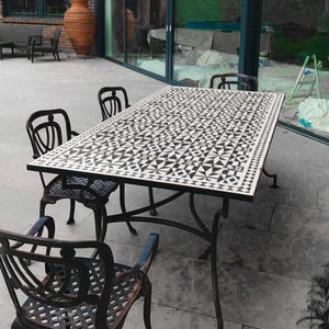 Puede incluir: Juego de comedor al aire libre con una mesa rectangular con una tapa de azulejos geométricos en blanco y negro. La mesa está sostenida por patas de hierro forjado y está rodeada por cuatro sillas a juego.
