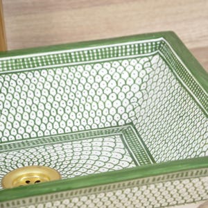 K&ouml;nnte beinhalten: Ein rechteckiges Keramikwaschbecken mit gr&uuml;nem Rand und wei&szlig;em Innenraum mit einem sich wiederholenden Muster aus gr&uuml;nen Tropfenformen. Das Waschbecken hat einen goldfarbenen Abfluss, ideal f&uuml;r ein Badezimmer.