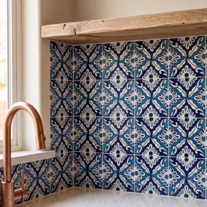 Könnte beinhalten: Küchenrückwand mit blau-weiß gemusterten Fliesen mit floralem Design. Die Fliesen sind in einem Raster angeordnet. Links ist ein Wasserhahn aus Kupfer zu sehen, und über den Fliesen befindet sich ein Holzregal. Die Fliesen sind ca. 15 cm x 15 cm groß.