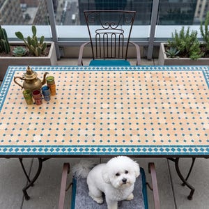 Puede incluir: Una mesa rectangular de exterior con una tapa de mosaico en beige y turquesa. La mesa está rodeada de sillas de metal con cojines turquesa. Una tetera de latón y vasos coloridos están sobre la mesa. Un perro blanco pequeño está sentado en una silla.