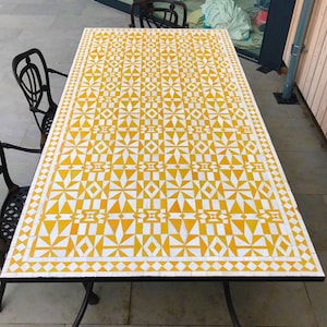 May include: Rectangular outdoor table with a white and yellow geometric tile pattern. The table has a black metal frame and legs. The table's dimensions are approximately 60 inches long and 30 inches wide.