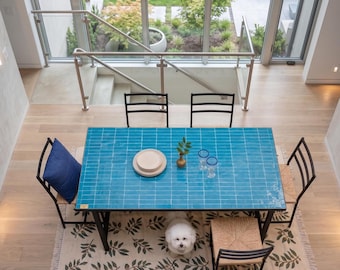 Pode incluir: Vista aérea de uma sala de jantar com uma mesa de azulejos turquesa, cadeiras pretas e um tapete estampado. A mesa está posta com pratos, copos e um pequeno vaso. Um cão branco está debaixo da mesa.