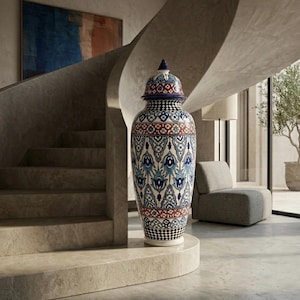 Può includere: Un grande vaso decorativo in ceramica con coperchio, caratterizzato da un motivo dettagliato in blu, rosso e bianco. Il vaso è posizionato su una base di pietra vicino a una scala a chiocciola. Il design include elementi floreali e geometrici.