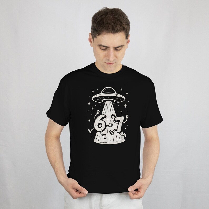 May include: Black t-shirt featuring a white graphic of a UFO beaming down the numbers 6 and 7, with arms and legs. The design includes stars and a simple, cartoonish style.