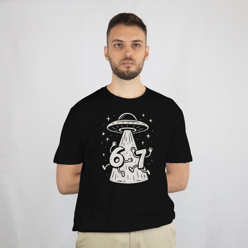 May include: Black t-shirt featuring a white graphic of a UFO beaming up the numbers 6 and 7. The design includes stars and a beam of light. The shirt is a crew neck style.