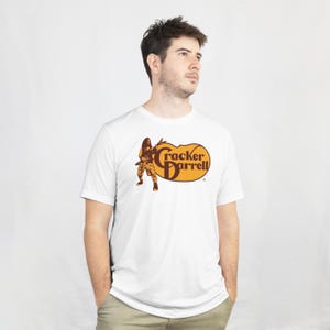 May include: White t-shirt featuring a brown and orange graphic of a person playing a guitar with the text "Cracker Darrell" in a stylized font. The shirt is made of a soft material and has short sleeves.