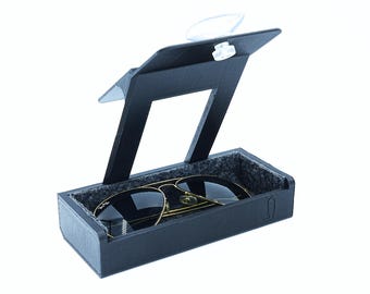 Op de afbeelding: Zwarte zonnebril met gouden montuur in een zwarte etui met een scharnierend deksel. De etui heeft een grijze viltvoering. Het deksel wordt opengehouden door een doorzichtig plastic onderdeel. De zonnebril is een klassieke pilotenbril.